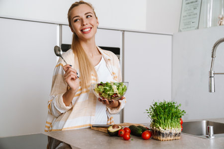 Joyful blonde woman smiling while preparing fresh salad at home kitchenの写真素材