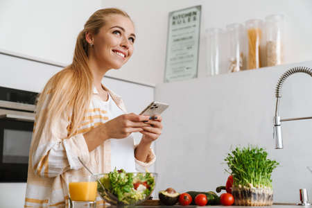 Portrait of a happy attractive woman using mobile phone while making a healthy salad in the kitchenの写真素材