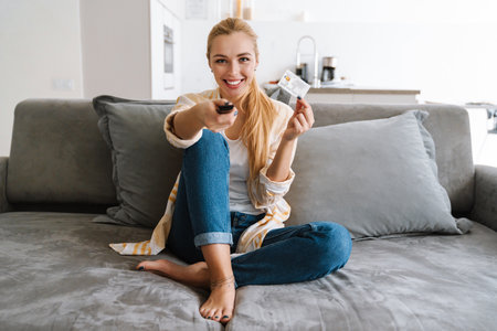 Happy woman holding credit card and watching tv with remote control at homeの写真素材