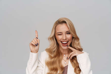 Beautiful happy girl in jacket winking and pointing finger upward isolated over grey backgroundの写真素材