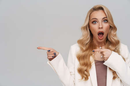 Beautiful shocked girl in jacket pointing fingers aside isolated over grey backgroundの写真素材