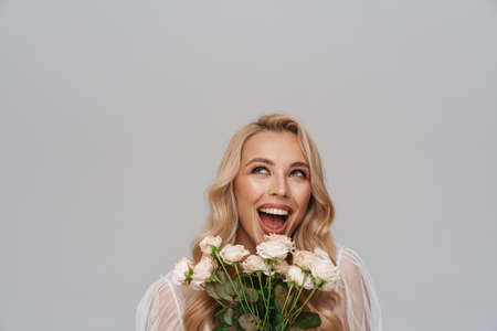 Beautiful smiling blonde haired young woman in dress standing isolated over blue background, holding rosesの写真素材