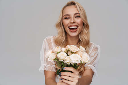 Beautiful smiling blonde haired young woman in dress standing isolated over blue background, holding rosesの写真素材