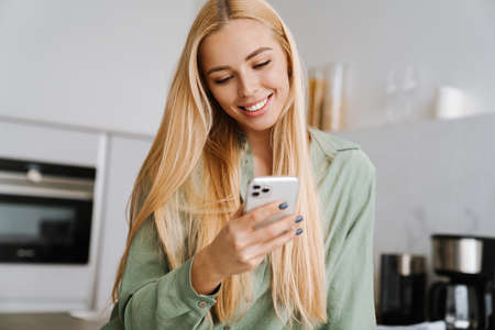 Happy blonde woman smiling and using cellphone at home kitchenの写真素材