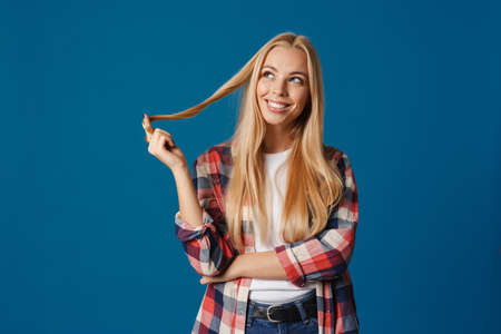 Blonde young happy woman smiling and playing with her hair isolated over blue backgroundの写真素材