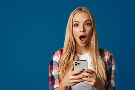 Blonde shock white woman exclaiming while using mobile phone isolated over blue backgroundの写真素材