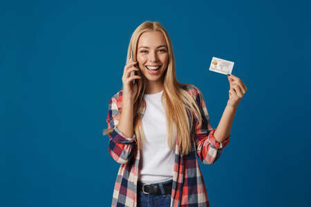 Blonde happy woman talking on cellphone and showing credit card isolated over blue backgroundの写真素材
