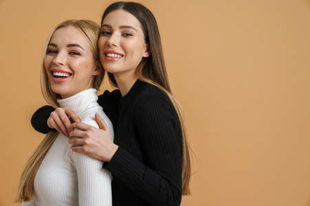 Two attractive smiling young sensual women embracing while standing isolated over beige backgroundの写真素材