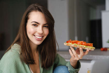 Young happy woman smiling while eating avocado toast at home kitchenの写真素材