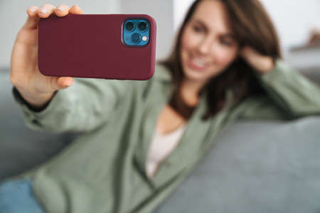 Happy brunette woman smiling and taking selfie photo on cellphone at homeの写真素材