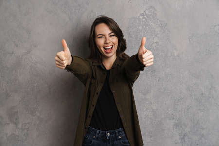 Portrait of happy smiling woman in casual wear standing isolated over gray background, giving thumbs upの写真素材