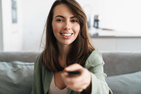 Happy young woman watching tv on sofa, holding remote controlの写真素材