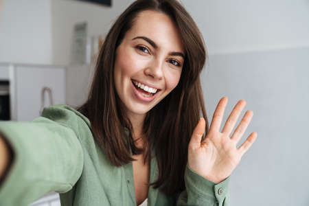 Young happy woman waving hand while taking selfie photo at home kitchen