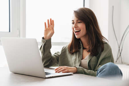 Smiling woman waving hand while taking video call on laptop at homeの写真素材
