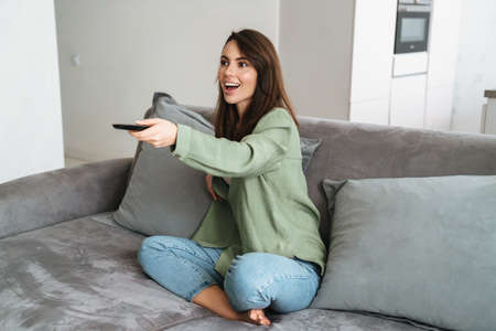 Happy young woman watching tv on sofa, holding remote controlの写真素材