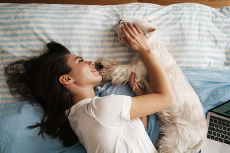 Happy white woman stroking her dog while lying on bed at homeの写真素材