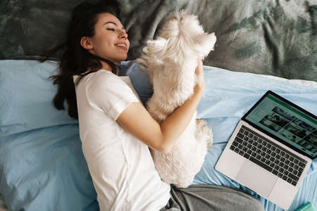 Happy white woman stroking her dog while lying on bed at homeの写真素材