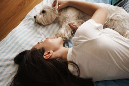 Happy white woman stroking her dog while lying on bed at homeの写真素材