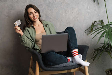 Smiling nice girl using laptop and credit card while sitting in chair indoors