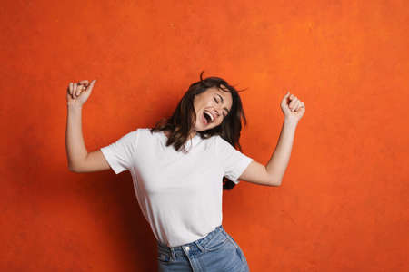 Excited beautiful girl singing and dancing on camera isolated over orange wall