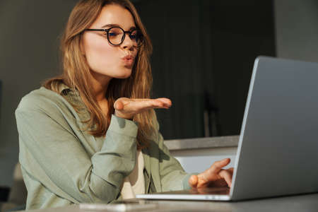 Happy nice woman blowing air kiss while working with laptop at homeの写真素材