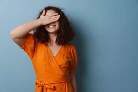 Happy redhead curly girl smiling and covering her eyes isolated over blue backgroundの写真素材