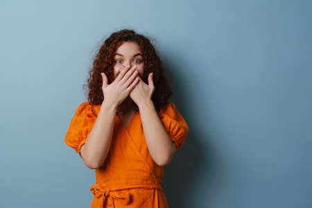 Redhead excited girl looking at camera and covering her moth isolated over blue background