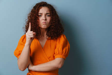 Irritated redhead curly girl wagging her finger at camera isolated over blue backgroundの写真素材