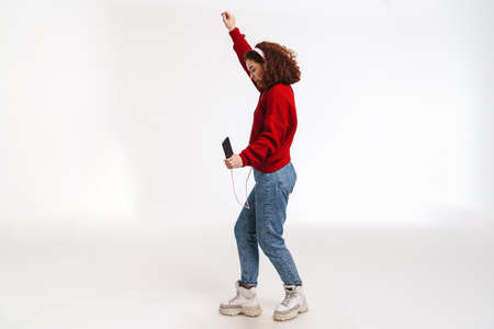 Joyful girl dancing while listening music with cellphone and headphones isolated over white...