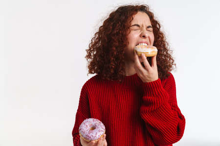 Excited ginger young woman grimacing while eating doughnuts isolated over white backgroundの写真素材