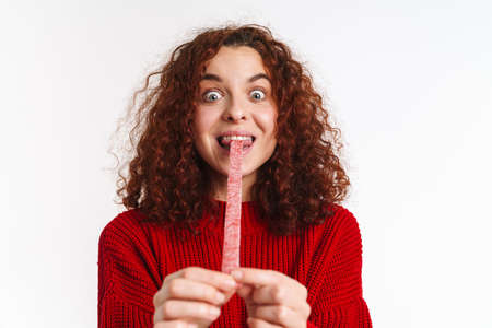 Joyful ginger curly girl grimacing while making fun with gummy isolated over white backgroundの写真素材