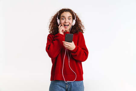 Excited ginger girl listening music with cellphone and headphones isolated over white background