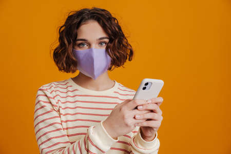 Young woman in facial medical mask using mobile phone isolated over yellow background