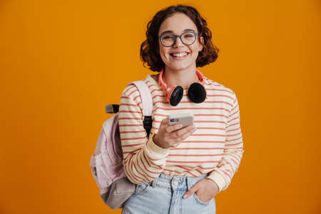 Cheerful young girl with backpack using mobile phone while standing isolated over yellow background