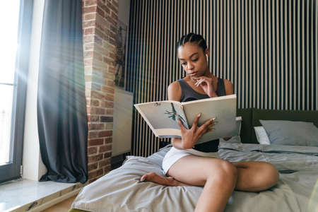 Focused african american girl in earphones reading book at bedroomの写真素材