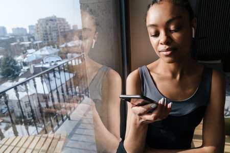 Happy african american girl in wireless earphones using mobile phone at home roomの写真素材