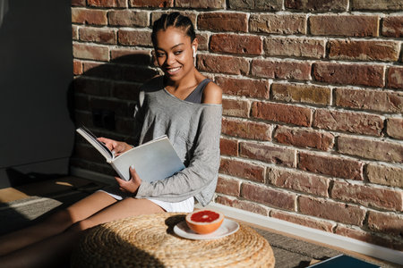 Smiling young african woman listening to music with earphones at home, reading a magazineの写真素材