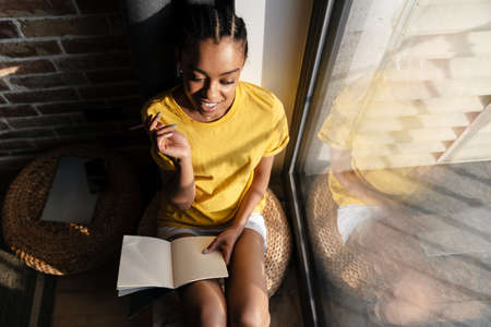 Happy african american girl in earphones writing down notes while sitting at the window at home