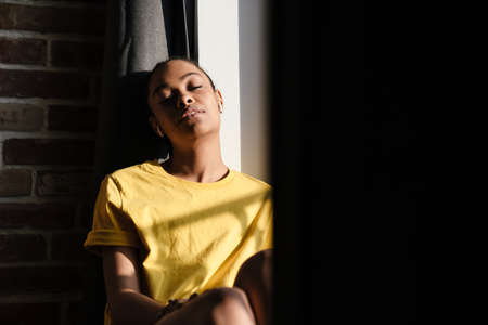 Relaxed african american girl listening music with earphones while sitting at window in homeの写真素材