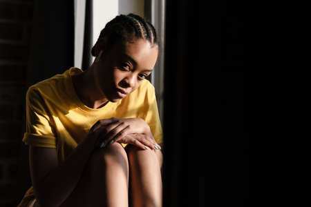 Attractive young african woman sitting on a windowsill at home, relaxing