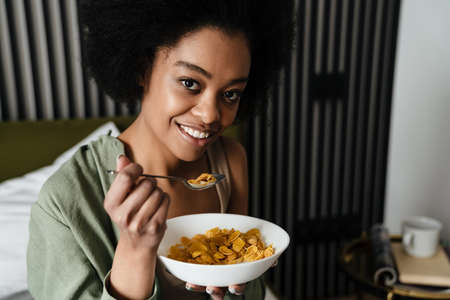 Smiling african american young woman eating breakfast while sitting in bedの写真素材