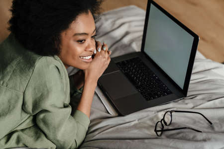Happy Mid aged african woman using laptop computer while laying in bed at homeの写真素材