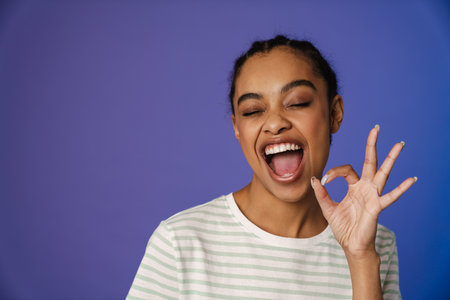 Young black woman in t-shirt laughing and gesturing ok sign isolated over blue background