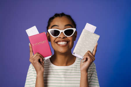 Happy young african woman showing passport and tickets over blue wall background