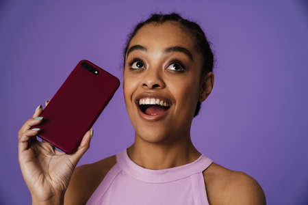 Young black woman smiling and holding mobile phone isolated over purple background