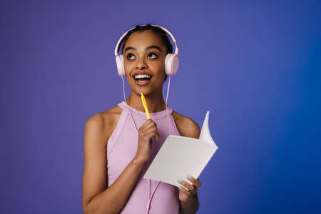 Close up of a smiling young african woman wearing headphones standing over blue wall, holding pen...