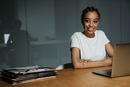 Black smiling woman working with laptop while sitting at table in officeの写真素材