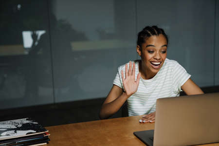 Black smiling woman gesturing and using laptop while working in office