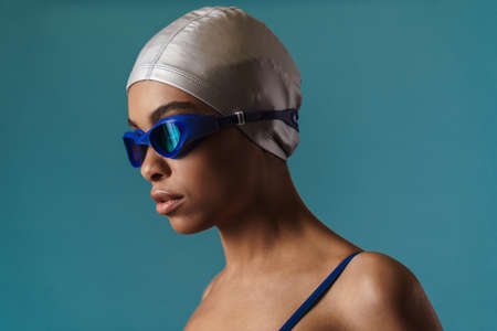 Close up portrait of a young african woman in swimsuit and swim hat standing over blue wallの写真素材