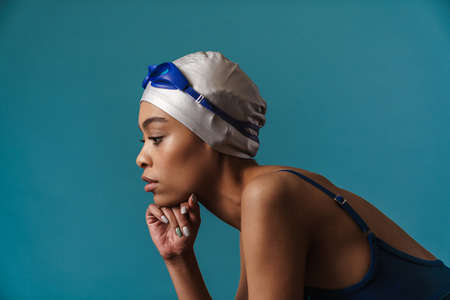 Young black swimmer in goggles posing and looking aside isolated over blue wallの写真素材